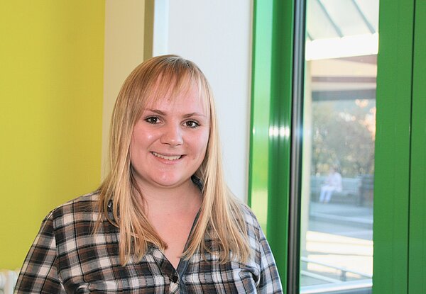 Eine junge Frau mit langen blonden Haaren lächelt vor einer hellen Wand und einem Fenster mit grünen Rahmen. Sie trägt ein kariertes Hemd und wirkt freundlich und zugänglich. Im Hintergrund sind unscharfe schemenhafte Figuren zu erkennen.