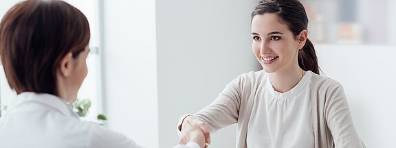 A young woman in a light cardigan smiles as she extends her hand for a handshake. She is seated across from another person, who is partially visible. The setting is bright and modern, suggesting a professional environment.