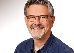 Christian Pfeiffer Ein lächelnder Mann mit grauem Haar und Brille steht vor einem hellen Hintergrund. Er trägt ein dunkelblaues Hemd und hat einen kurzen, gepflegten Bart. Der Ausdruck auf seinem Gesicht vermittelt Freundlichkeit und Offenheit.