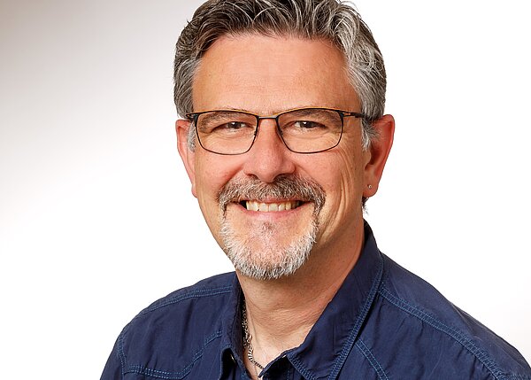 Ein lächelnder Mann mit grauem Haar und Brille steht vor einem hellen Hintergrund. Er trägt ein dunkelblaues Hemd und hat einen kurzen, gepflegten Bart. Der Ausdruck auf seinem Gesicht vermittelt Freundlichkeit und Offenheit.