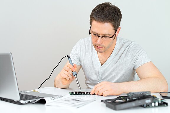 A man in a gray shirt is soldering components on a circuit board, focusing intently on his work. A laptop sits on the table alongside various electronic parts and a manual. The setting suggests a hands-on electronics project or repair.