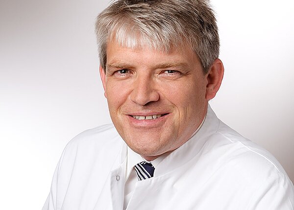 Ein lächelnder Arzt in weißem Kittel steht vor einem hellen Hintergrund. Sein Haar ist kurz und hell, und er trägt ein gestreiftes Hemd unter dem Kittel. Sein Gesichtsausdruck vermittelt Freundlichkeit und Professionalität.