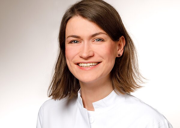 Ein Lächeln einer Frau mit schulterlangen braunen Haaren, die eine weiße Arbeitskleidung trägt. Der Hintergrund ist hell und neutral. Die Frau strahlt Freundlichkeit und Offenheit aus.