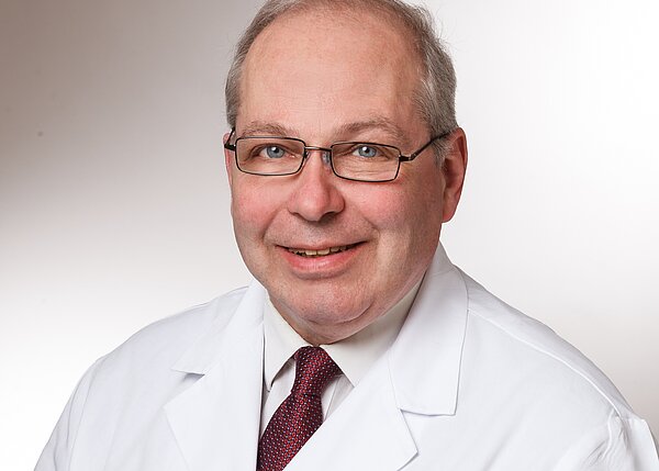 Ein lächelnder Arzt trägt einen weißen Kittel und eine Krawatte. Er hat Brille und graue Haare. Der Hintergrund ist neutral und hell, was auf eine professionelle Atmosphäre hinweist.