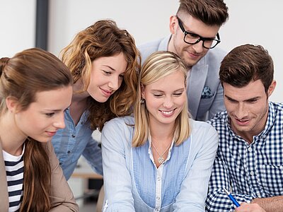 Sechs Personen stehen um einen Tisch und schauen konzentriert auf etwas, das sich dort befindet. Sie scheinen an einem Projekt oder einer Diskussion zu arbeiten, wobei ihre Mienen interessiert und engagiert wirken. Ausbildungsmoeglichkeiten_90978297.jpg